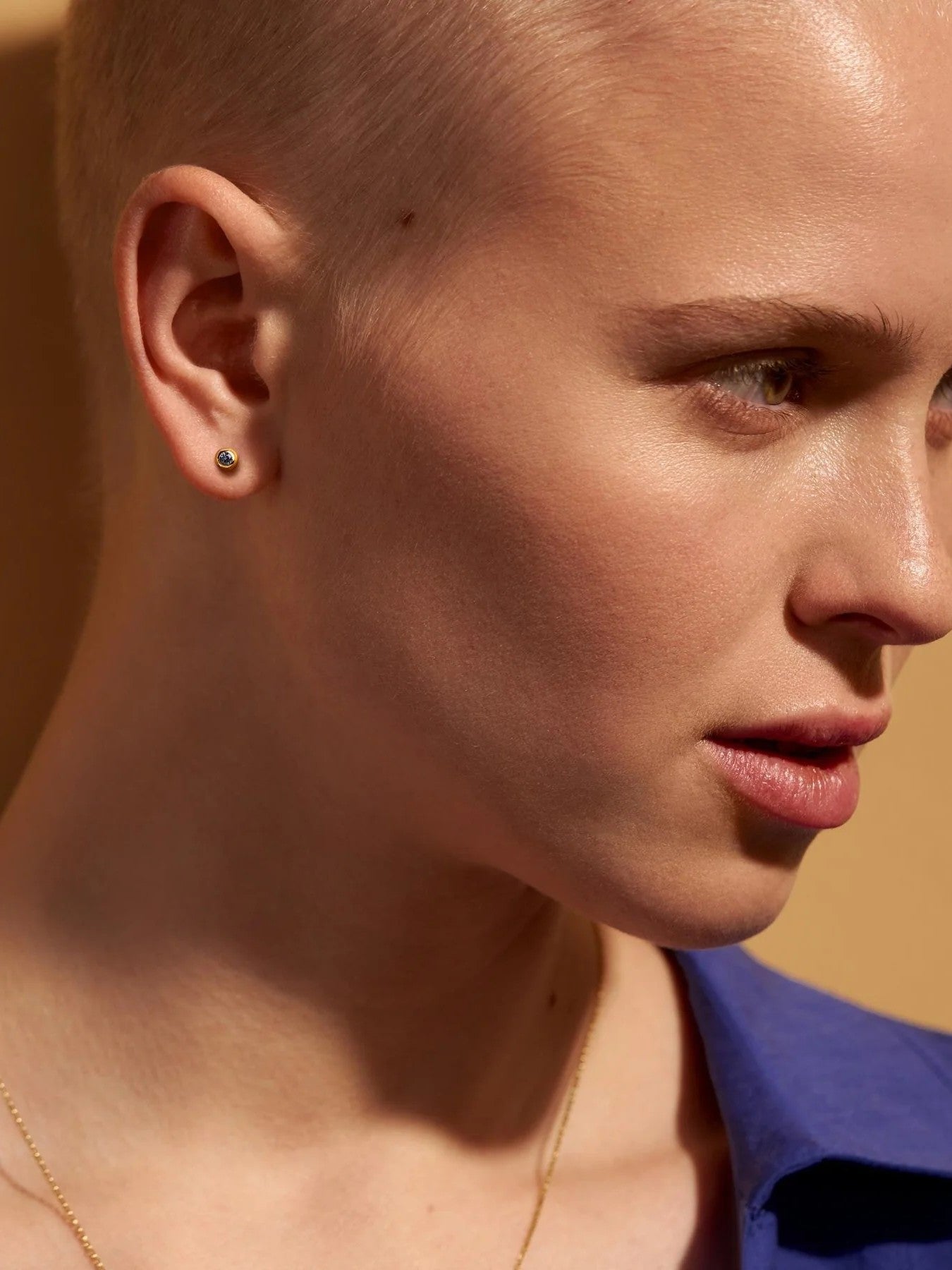 Model with a shaved head wearing a gold earring against a warm-toned background.