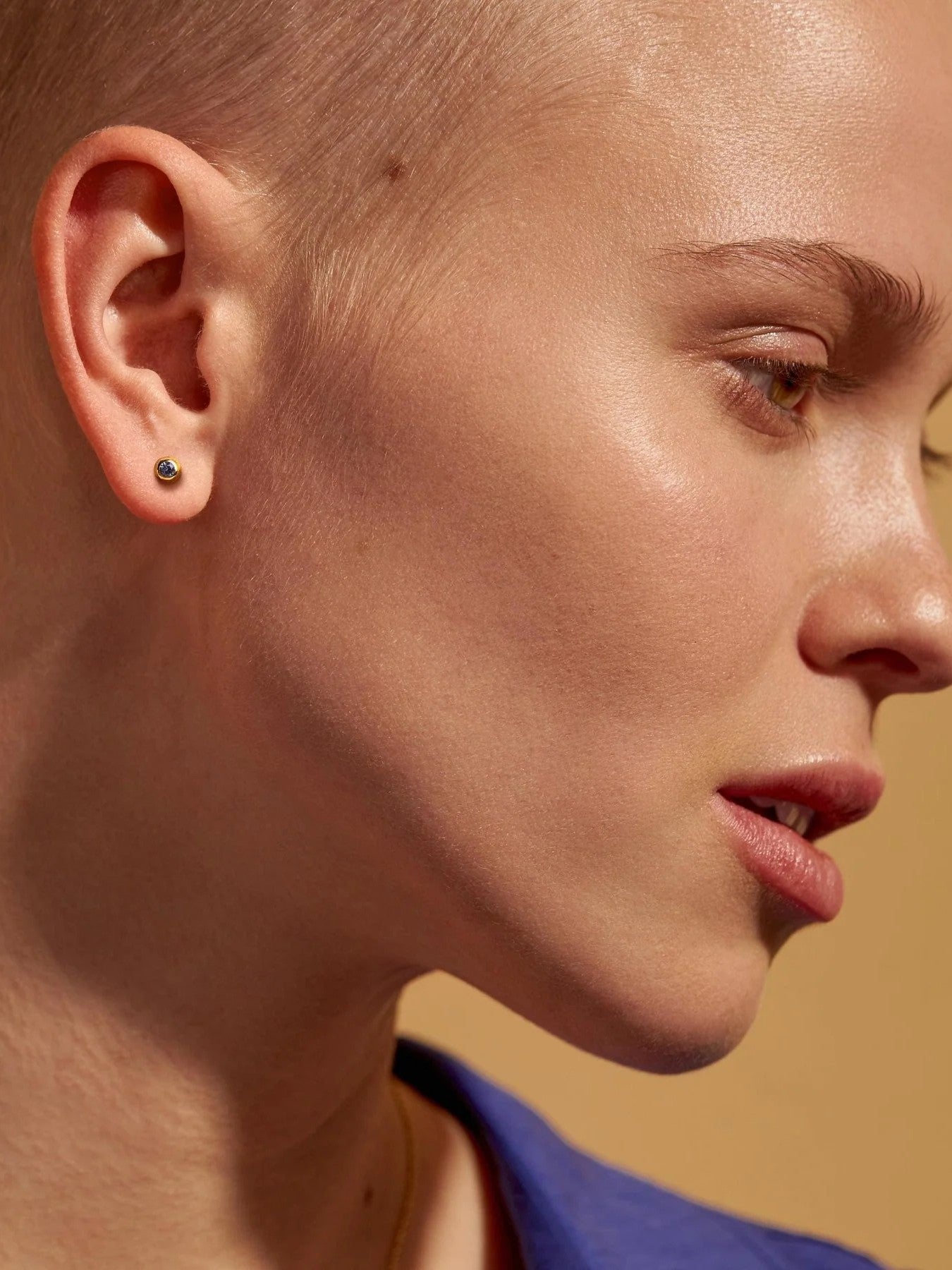 Profile portrait of a model with a shaved head wearing a gold earring with a dark stone.