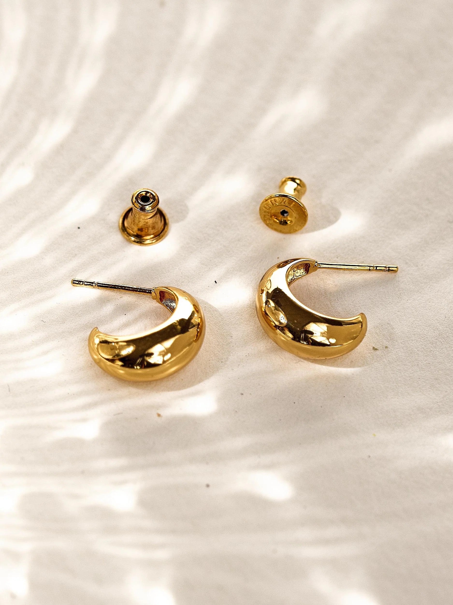Gold plated crescent hoop earrings with matching backs, photographed from the front on a softly textured background.