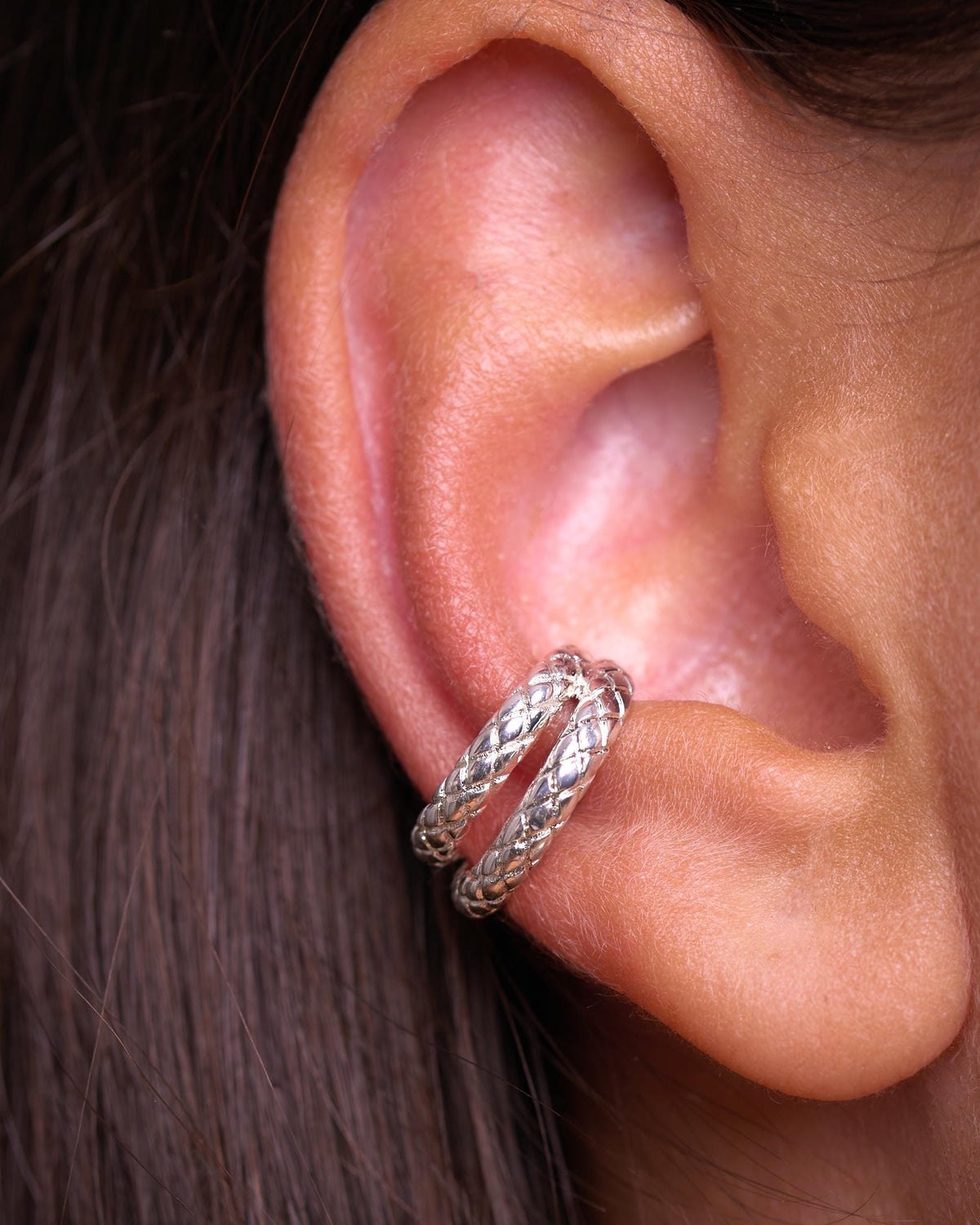 Silver snake ear cuff worn on the middle of the ear, close-up of a woman's ear with dark hair. Double band cuff with a textured snake scale pattern, no piercing needed.