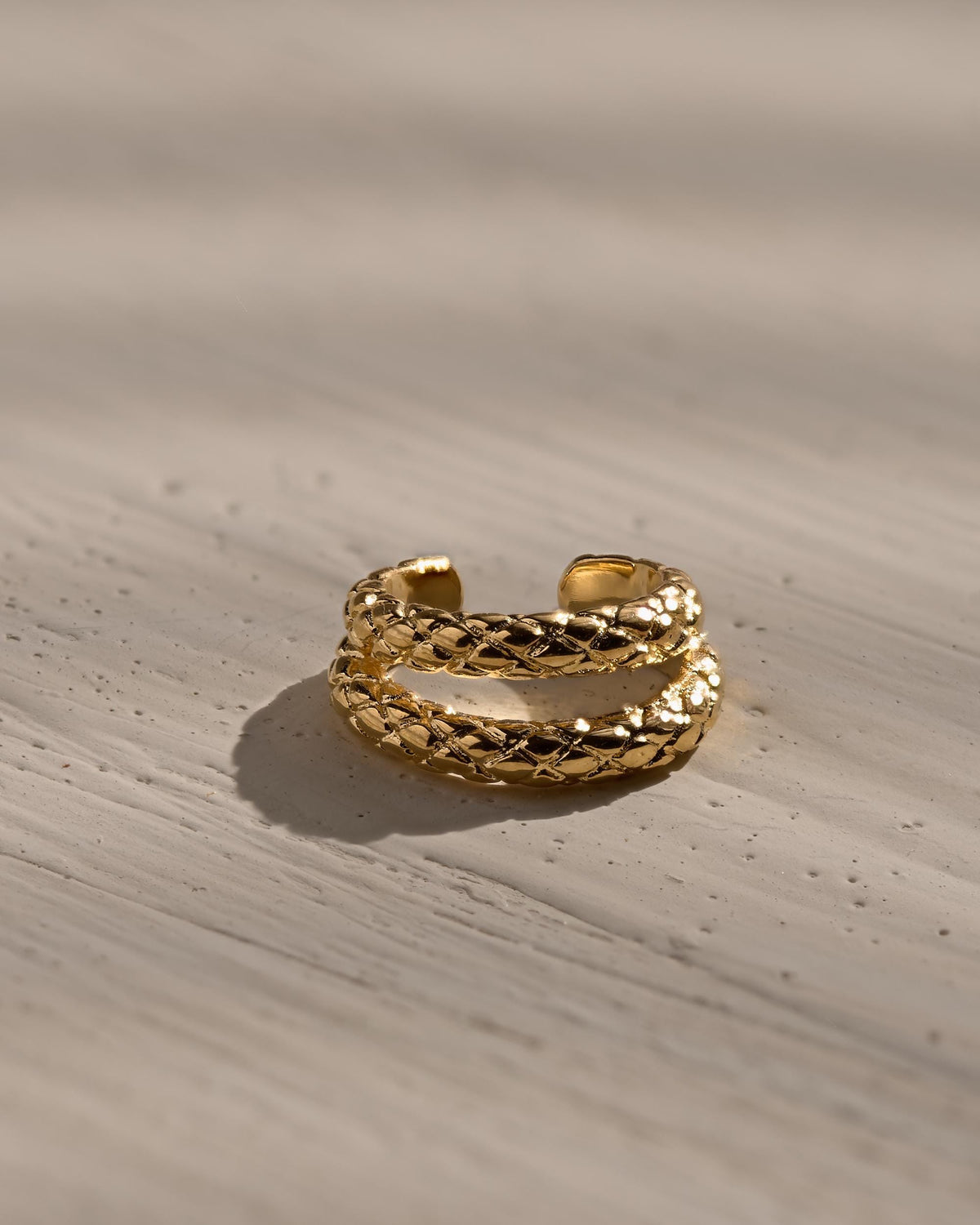 A gold-toned open ring featuring a double-band design with a textured, quilted, or woven-like pattern across the surface. The ring has a polished metallic finish and an open gap at the back, allowing for potential adjustment in fit.