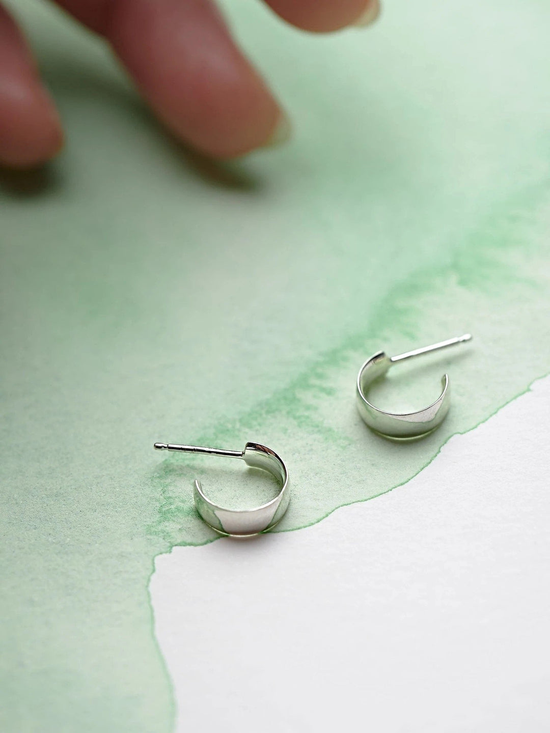 Pair of petite sterling silver hoop earrings on textured green and white background, hand reaching in.