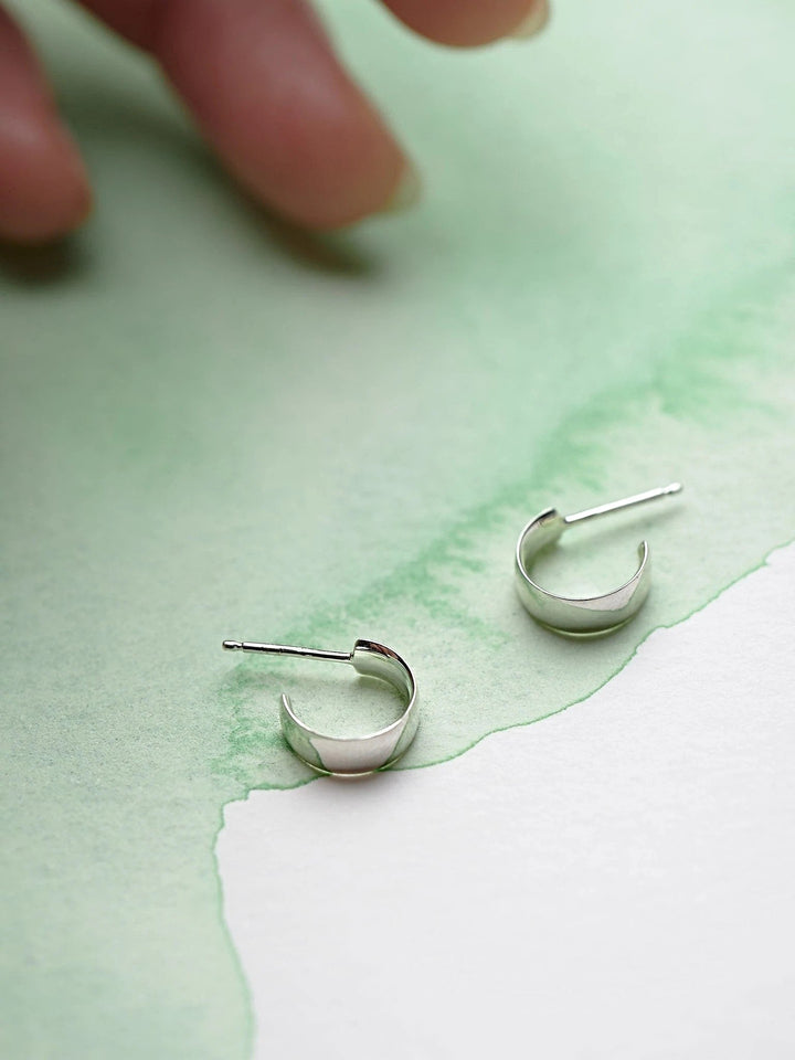 Pair of petite sterling silver hoop earrings on textured green and white background, hand reaching in.