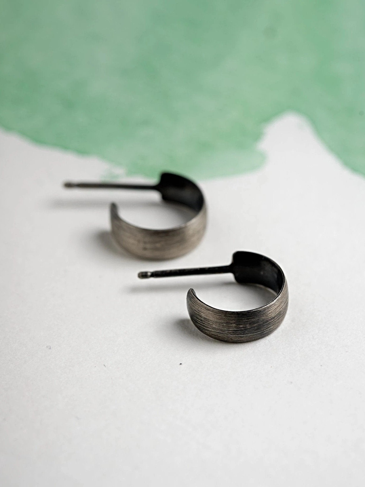 Side view of tiny silver oxide hoop earrings on green textured paper.