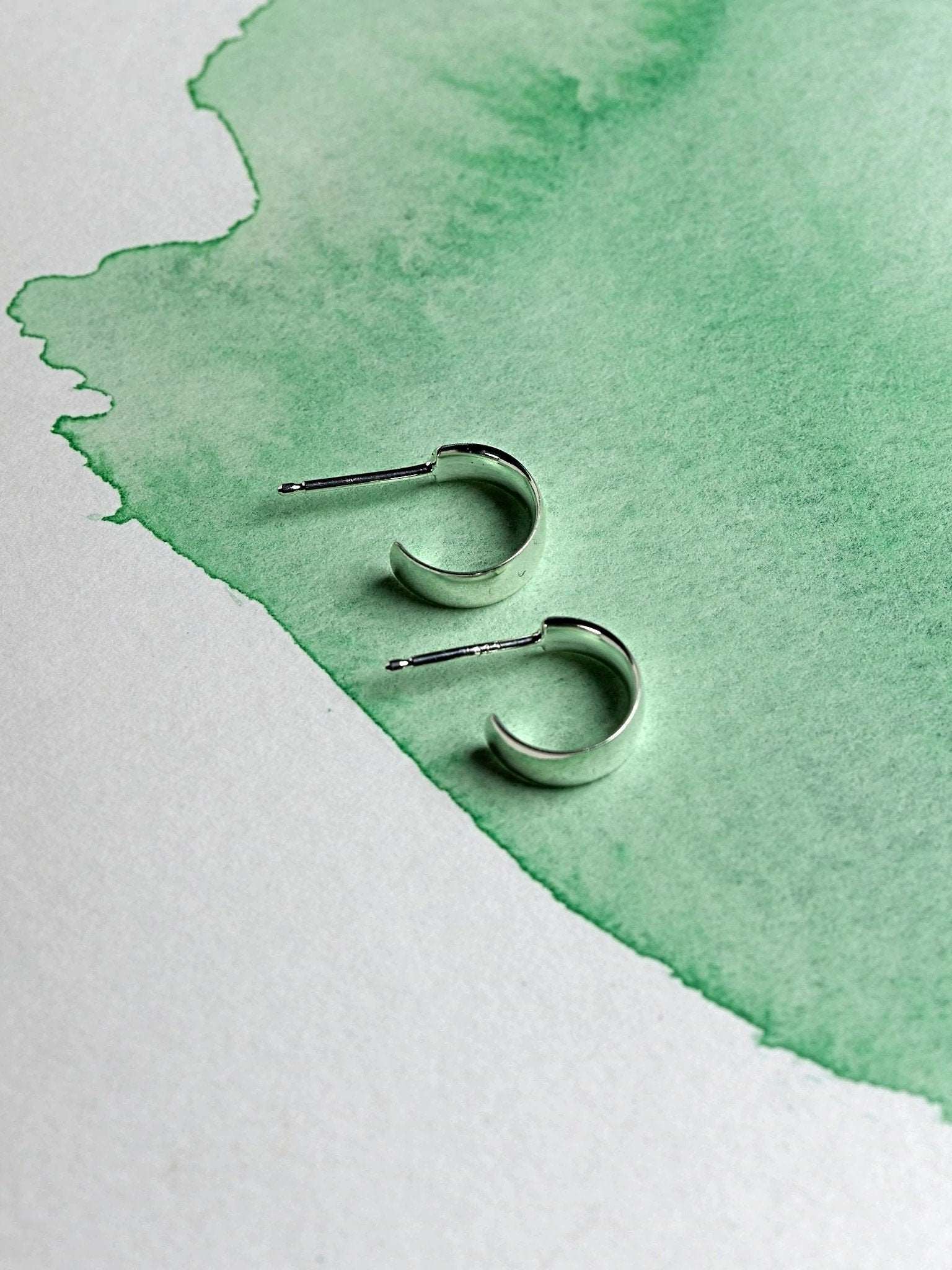Minimalist sterling silver hoop earrings flat-lay on watercolor green paper.