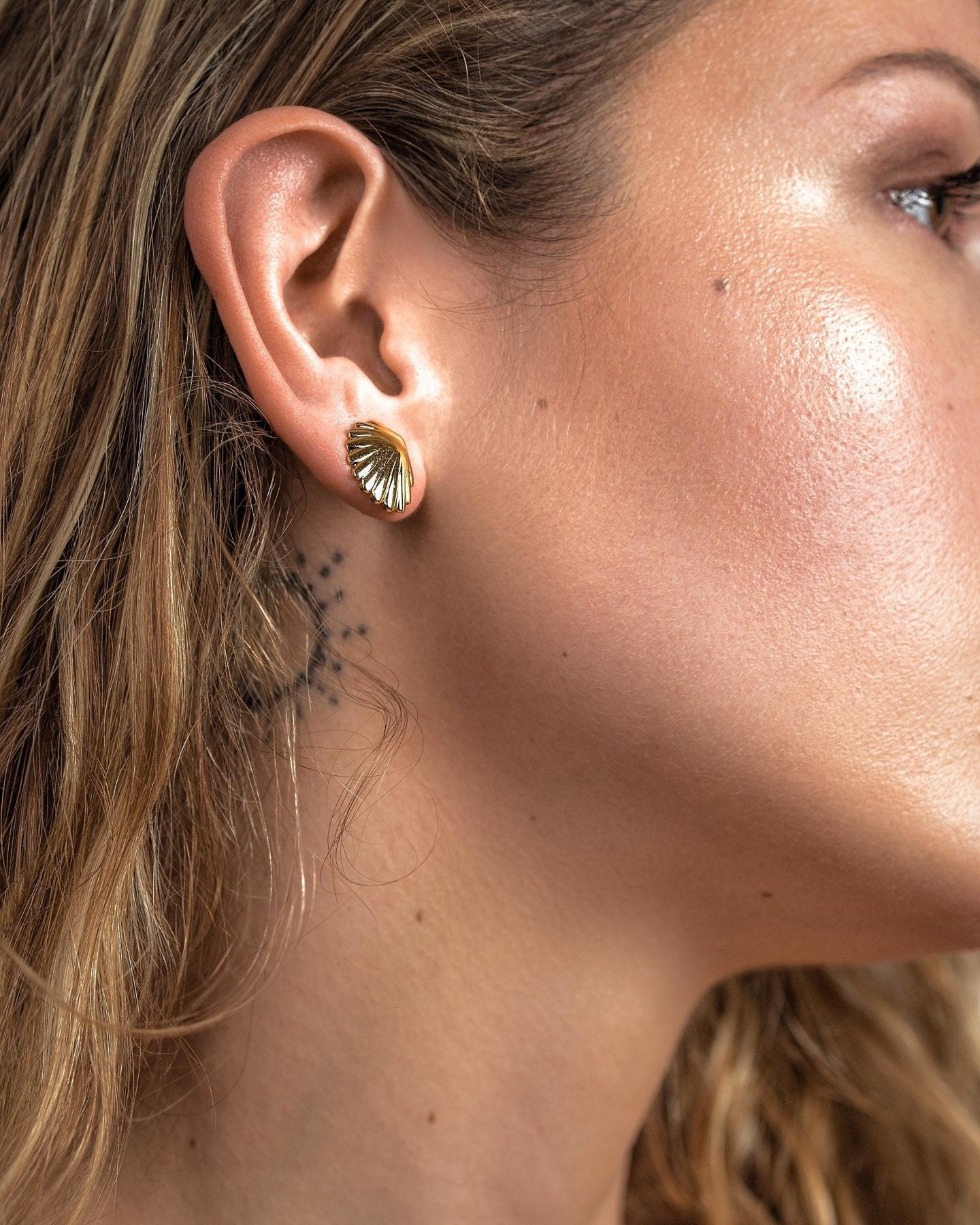 Close-up of a model wearing a small gold scallop shell stud earring against the earlobe.