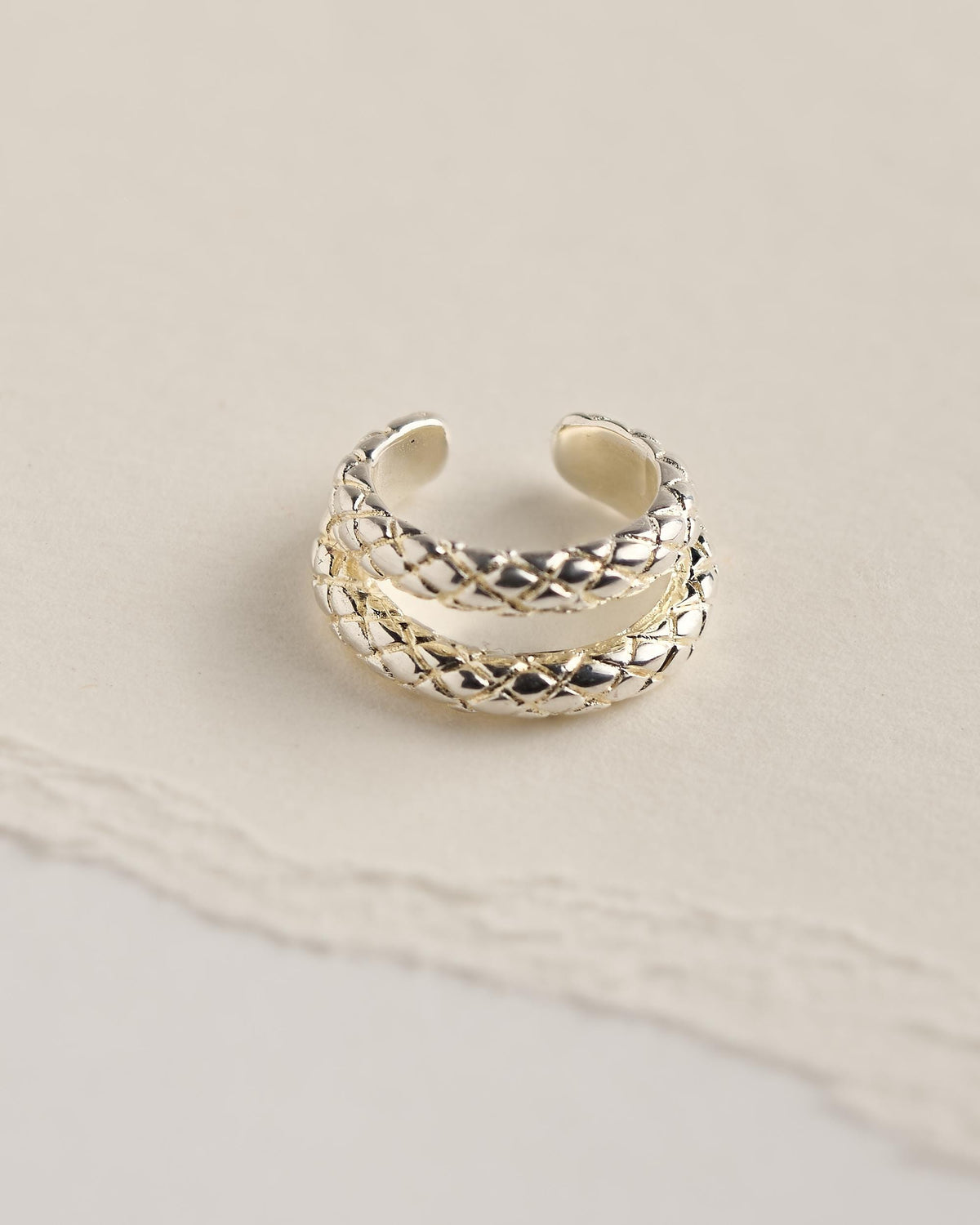Sterling silver plated brass snake ear cuff with two bands of engraved snake-scale texture, seen from above on cream torn paper.