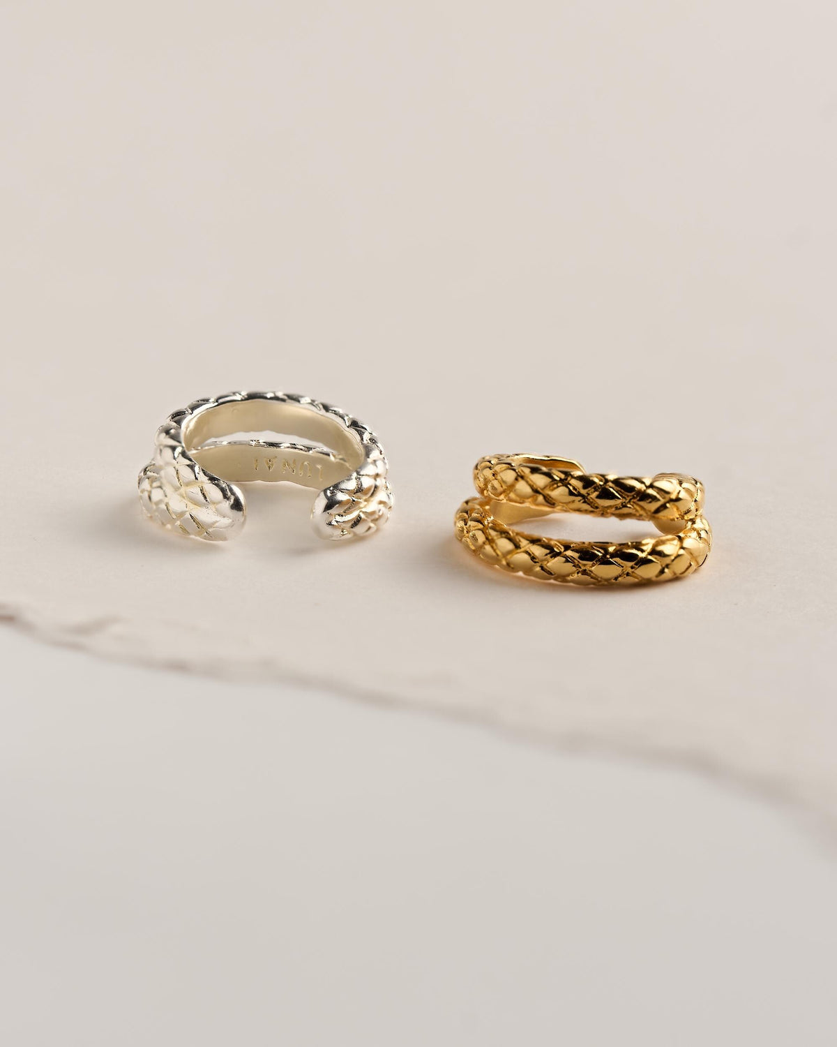 Two snake ear cuffs in sterling silver plated brass and 24k gold plated brass, with engraved snake-scale texture, on cream paper.