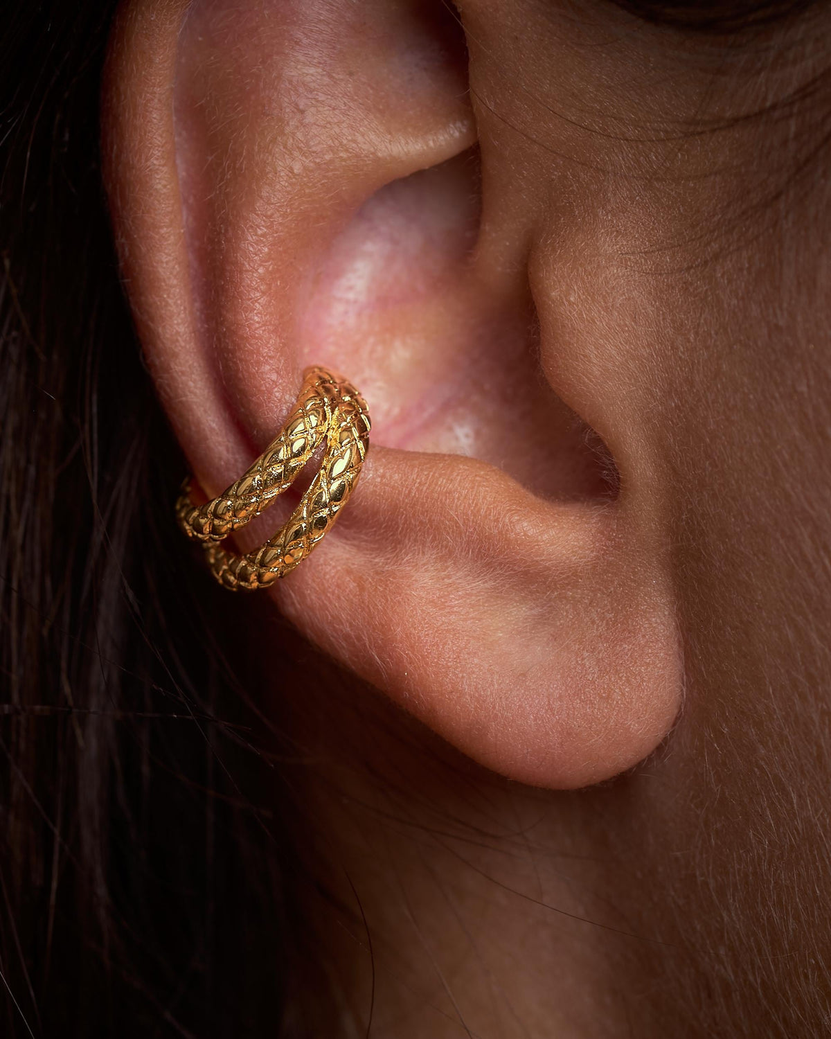 Close-up of an ear wearing a 24k gold plated brass snake ear cuff on the conch, with two bands of engraved snake-scale texture.
