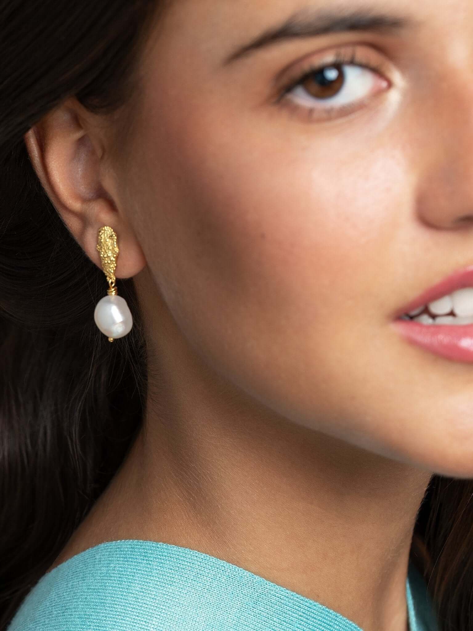 Model wearing baroque pearl earrings, front view with soft smile and blue top.